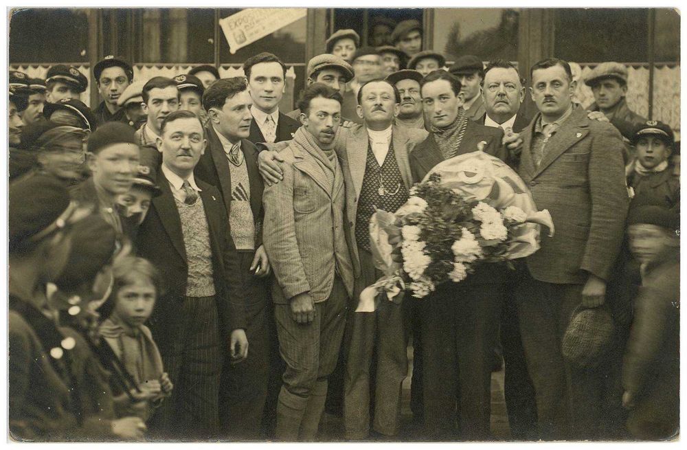 Photographie en noir et blanc, on voit au centre de l'image un homme poser fièrement ses bras sur les épaules de deux jeunes hommes de parts et d'autres de lui. L'un deux tient une gerbe florale dans ses mains. Autour, des officiels posent et au premier plan, des enfants flous regardent l'objectif.