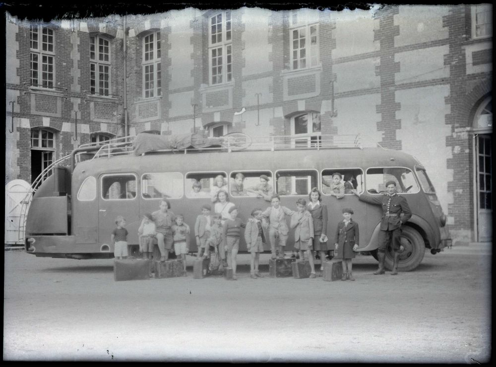 Photographie en noir et blanc d'un bus garé en longueur devant une habitation en brique marquant un angle. Devant et à l'intérieur, posent une vingtaine de personnes : des enfants, des instructrices et un homme qui semble être le conducteur du bus, accoudé à ce-dernier. À leurs pieds, des valises. Le bus possède une rampe qui permet de stocker des bagages sur le toit, ainsi qu'une remorque incurvée.