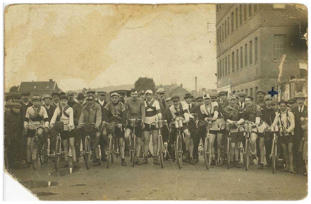 Carte postale jaunie de la ligne de départ des cyclistes, ils se tiennent à côté de leur vélo, collés les uns aux autres sur un chemin en terre battue. Ce sont tous des jeunes hommes, ils portent des shorts et des couvre-chefs.