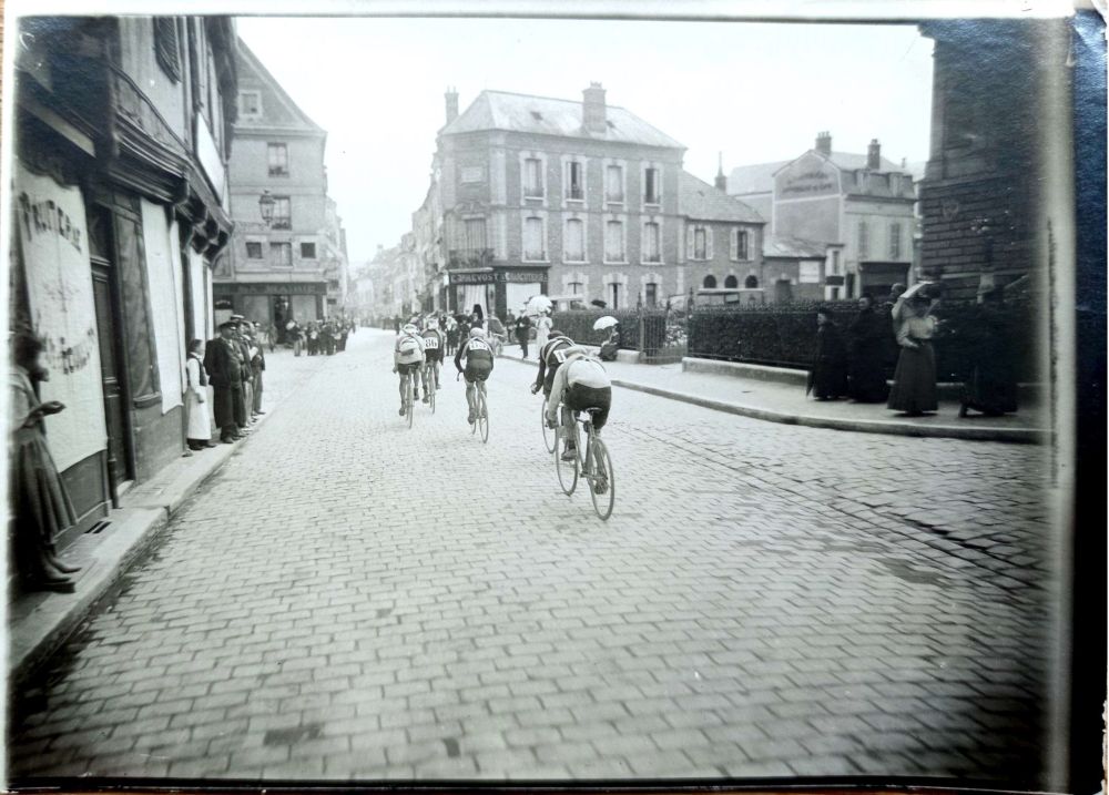 Photographie en noir et blanc d'une rue pavée où l'on voit quelques cyclistes de dos et des passants, hommes, femmes et enfants les regardant sur les trottoirs.