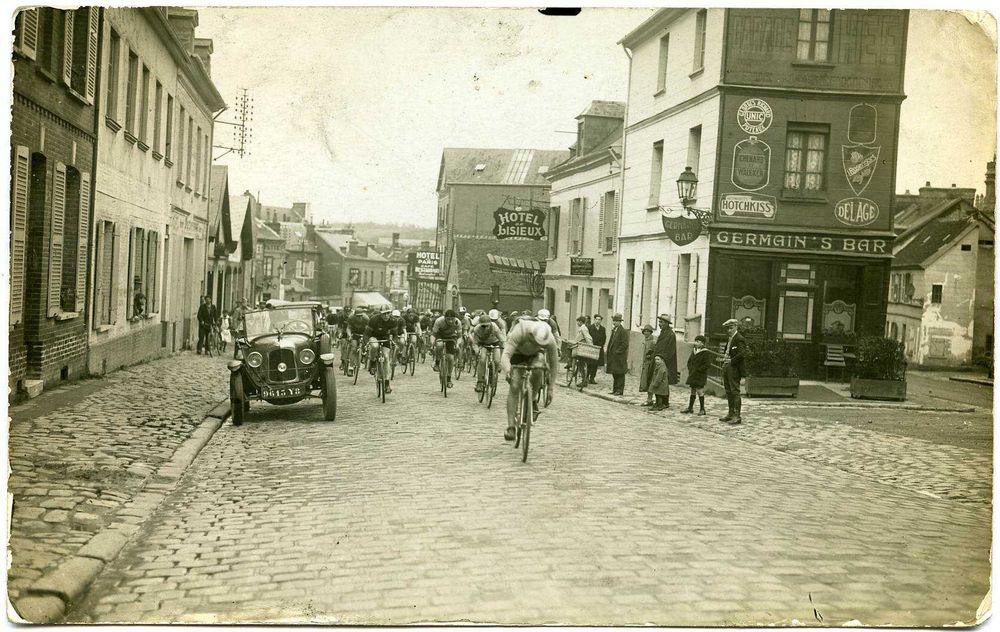 Carte postale jaunie où l'on voit le peloton de tête de face remonter une ruelle pavée, devant un bar nommé "Germain's bar", une voiture ancienne suit les cyclistes, acclamés par quelques personnes sur le trottoir à proximité du bar.