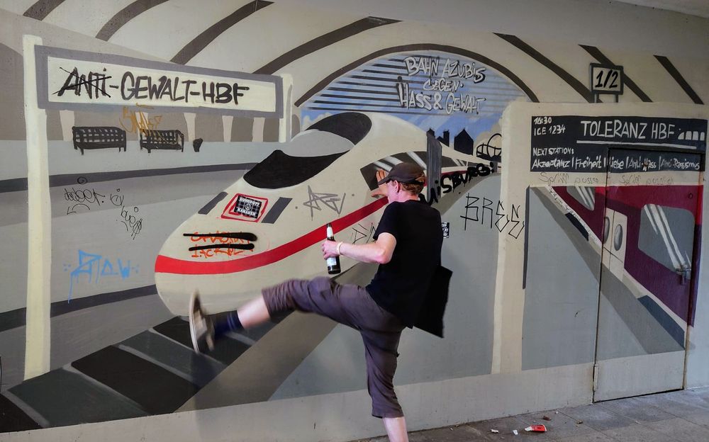 Eine Wand in einer Bahnunterführung, großflächig mit einem Wandbild bemalt, das einen Bahnsteig im Bahnhof zeigt mit einem einlaufenden ICE und Schildern, auf denen steht "Anti-Gewalt-Bahnhof" und "Toleranz-Hbf - Next Station: Akzeptanz / Freiheit / Liebe / Anti-Hass / Antirassismus". Vor dem Bild hampelt eine Person herum mit Bierflasche in der Hand und tut so, als würde sie nach dem (gemalten) Zug treten. 