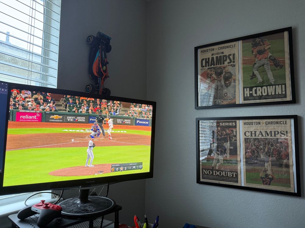 A photo of Sean’s second monitor with the Astros vs Mets playing, and his framed front pages of the Houston Chronicle from Houston’s two championships hanging to the right.