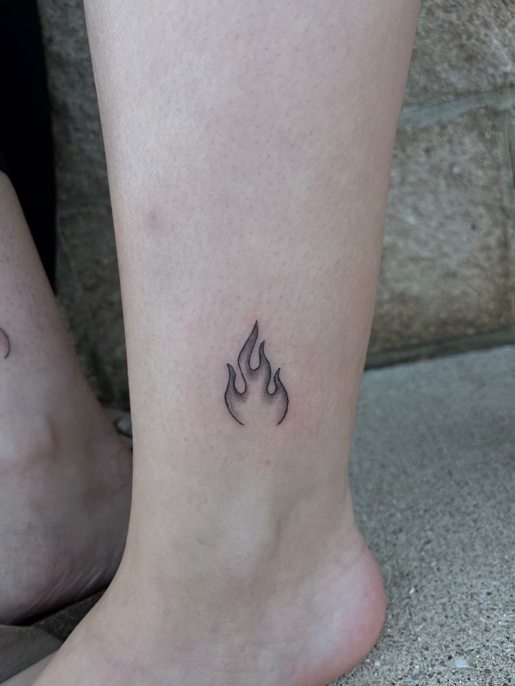 A small black and grey tattoo of a flame, placed on the outer ankle. 