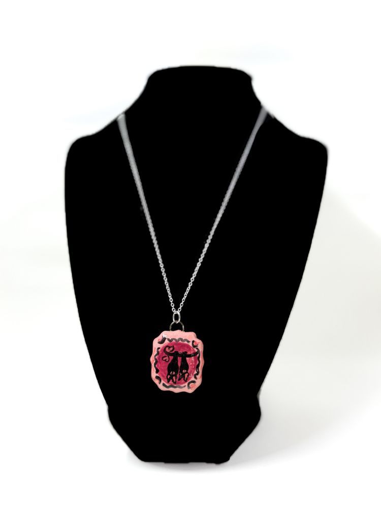 A black stand holds a necklace on a silver chain. The necklace pendant is painted pink, with two bat silhouettes hanging upside down from a black branch with heart-shaped accents. 