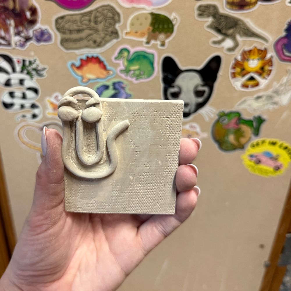 A hand holding up a ceramic tile with a sculpted “Clippy” paperclip