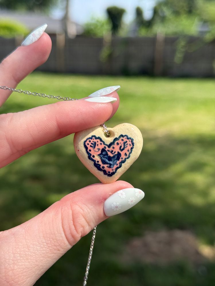 A hand holding up a heart shaped pendant necklace. The necklace is painted with a delicate, vintage design featuring a chicken silhouette