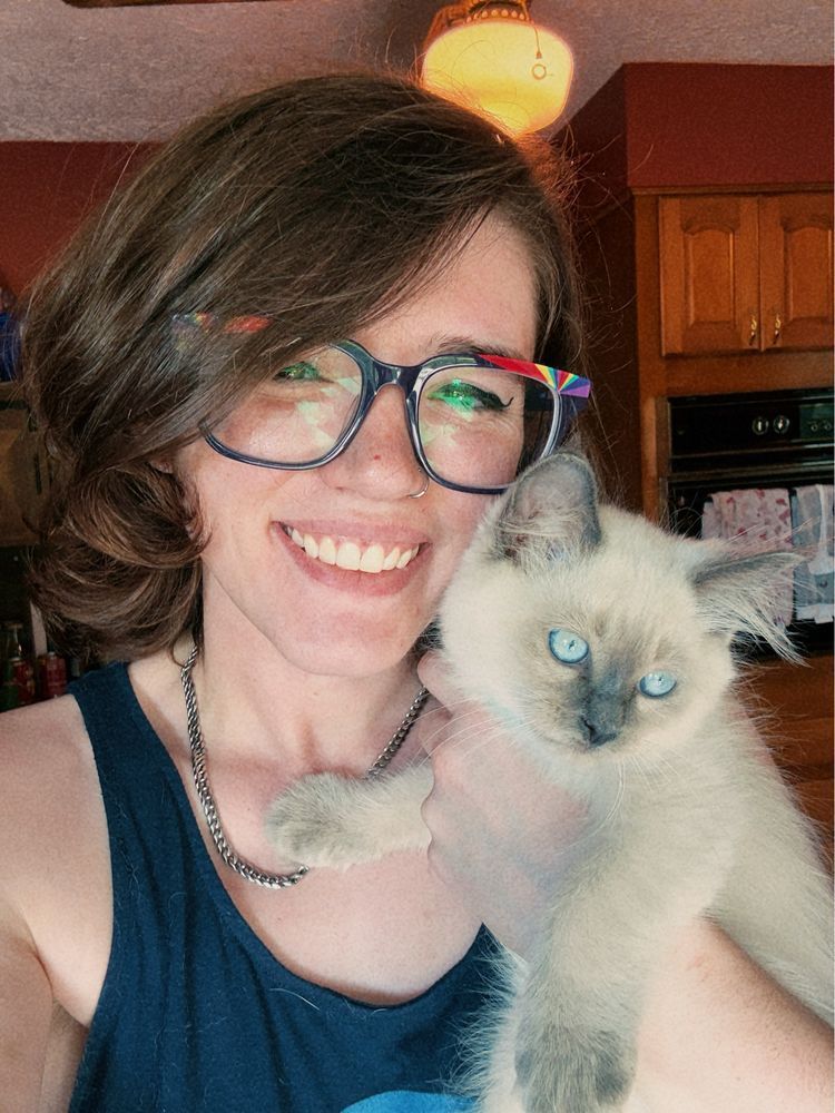 A white woman with brown hair and glasses smiling holding a white and brown kitten with bright blue eyes. 