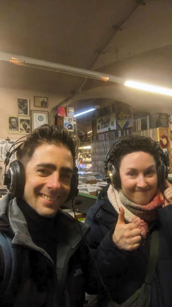 Me taking a selfie of me and my wife wearing headphones on Alt Neu. We're smiling and she's giving a thumbs up. (The listening booth was purpose built for the film and is not part of the record store).