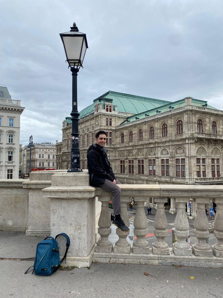 Me straddling a stone barrier at the top of the Albertina art museum. In the background is the Vienna State Opera House.