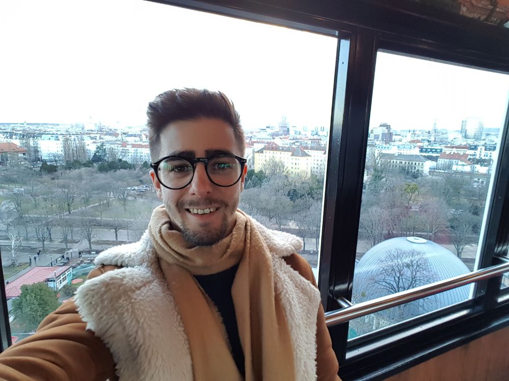 Me taking a selfie on the Ferris wheel in Prater amusement park. A window is behind me with a panoramic view of Vienna.