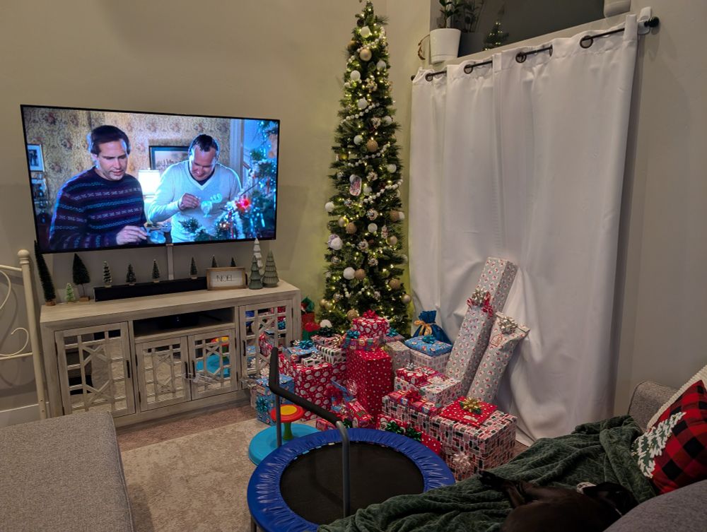 A pile of presents in front of a Christmas tree. A TV on the wall is playing National Lampoon's Christmas Vacation.