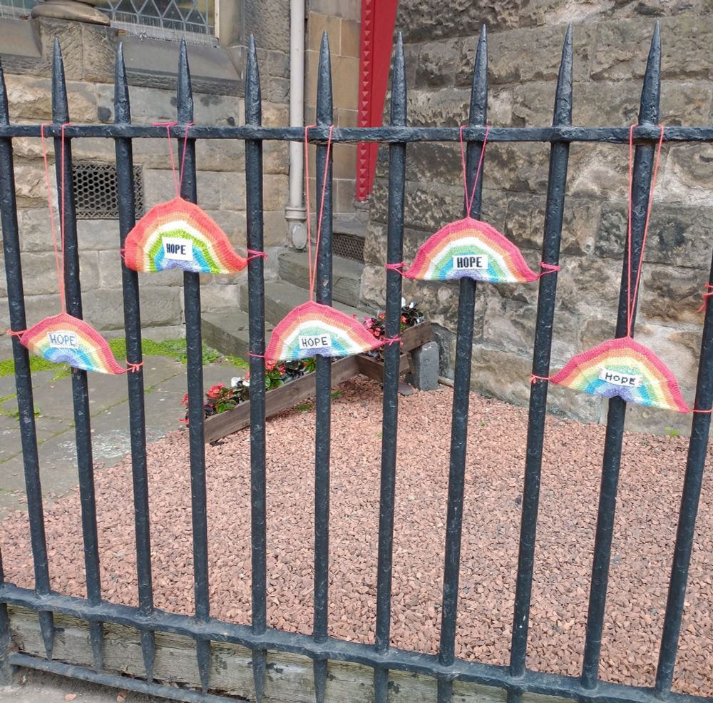 Five small knitted rainbows, each with the word 'HOPE' added at the centre, hanging from black-painted railings in front of a stone building.