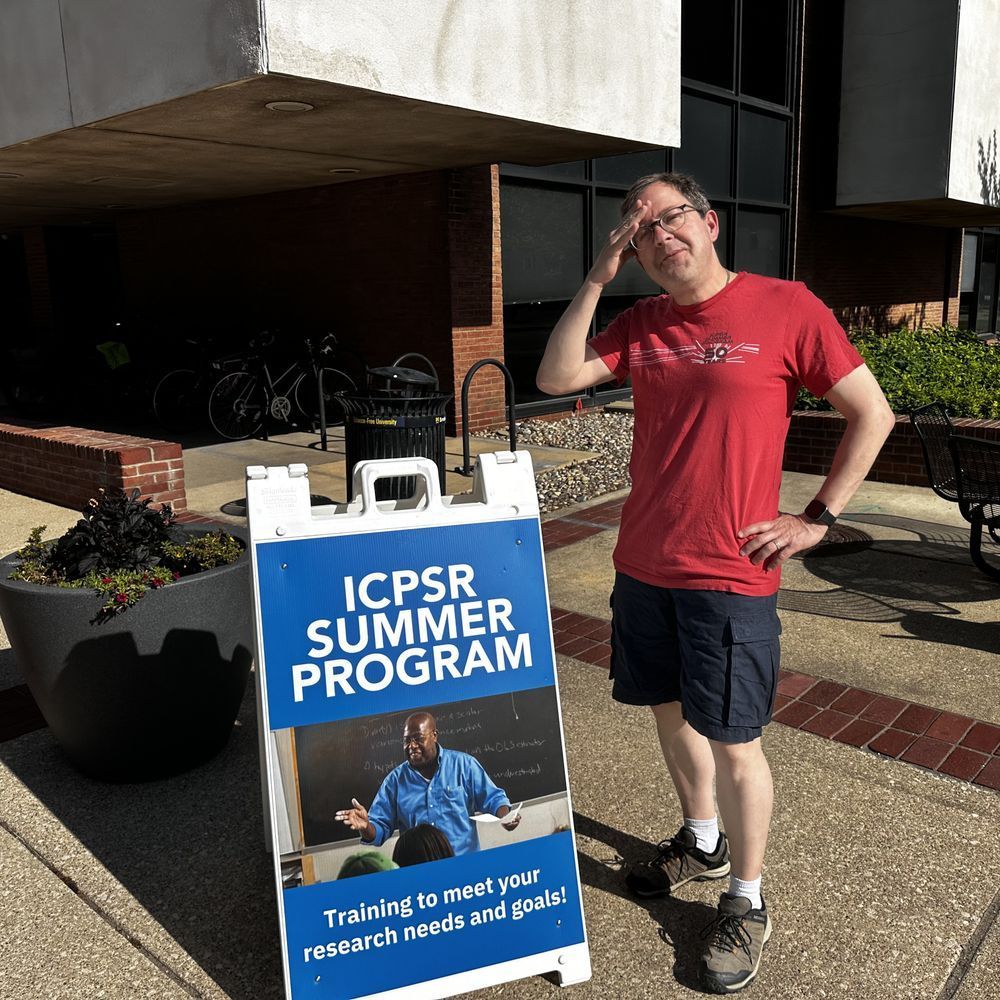 A man wiping sweat from his brow as he poses next to a Summer Program sign