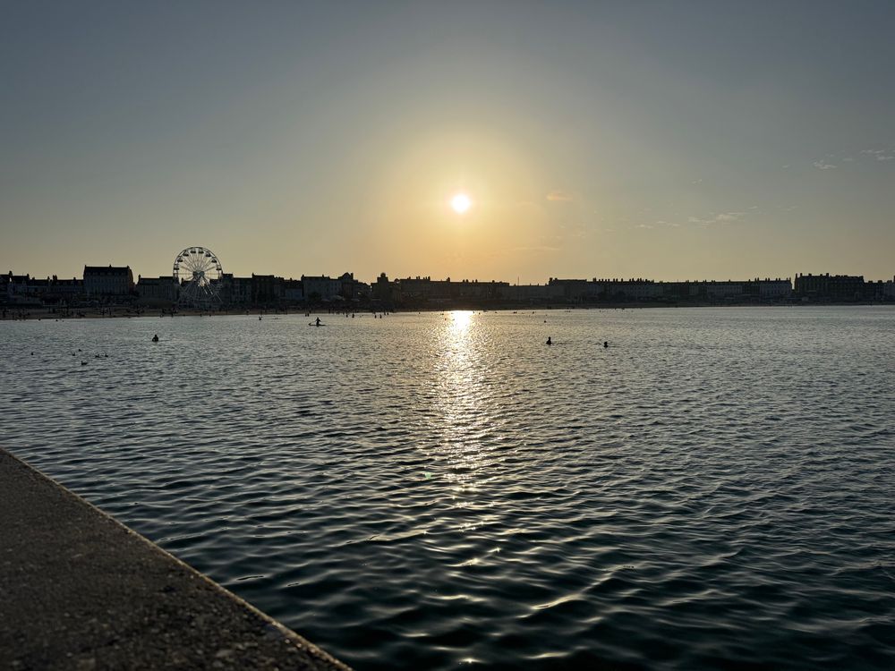 Panorama photo taken from Weymouth pier of the Sun setting over the beach, casting a reflection on the water. In silhouette you can make out dozens of people still swimming or paddle boating in the shallow water, while in the distance a funfair keeps people busy.