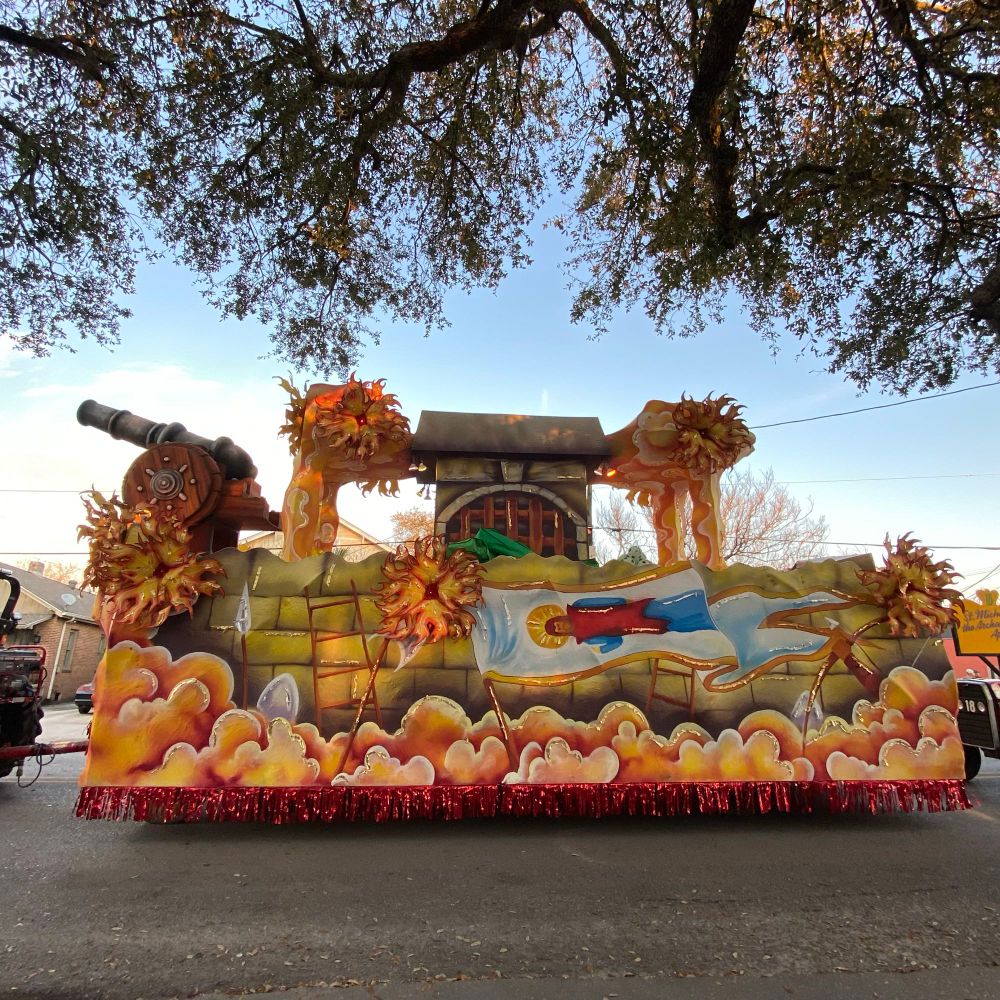 Ornate Mardi Gras float with (paper mache) cannon at the front and large yellow flowers edged in red. Bottom is edged with orange clouds and a skirt of red foil streamers. Oak tree branches fill the top of the frame.