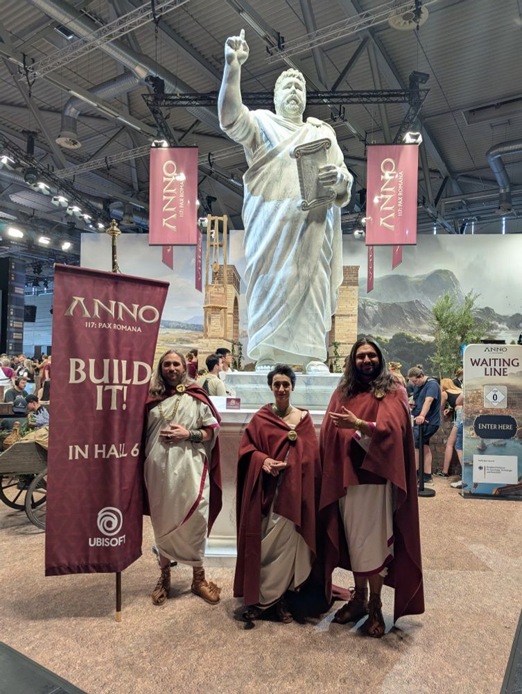 Three people dressed in ancient Roman costumes in front of a large but fake Roman statue on top of a plinth advertising the game Anno 117