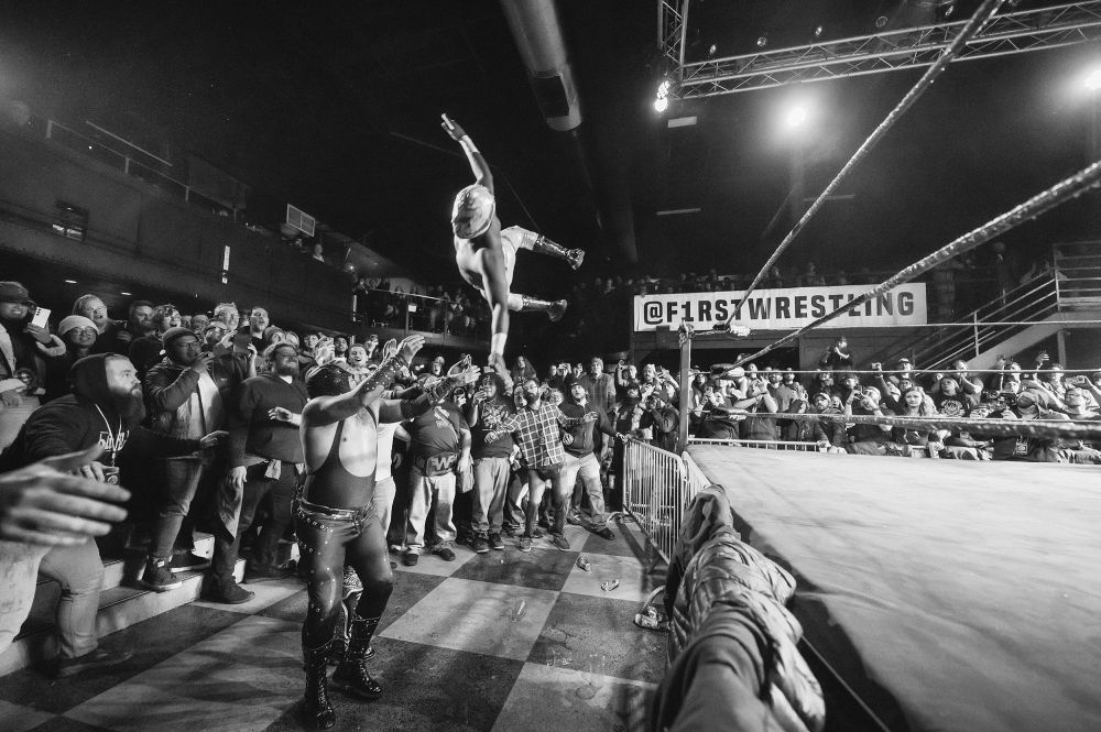 Black and white photograph of luchador wrestlers in action. One is jumping from the ropes of the ring while the other is in the middle of the crowd, ready to catch him. 