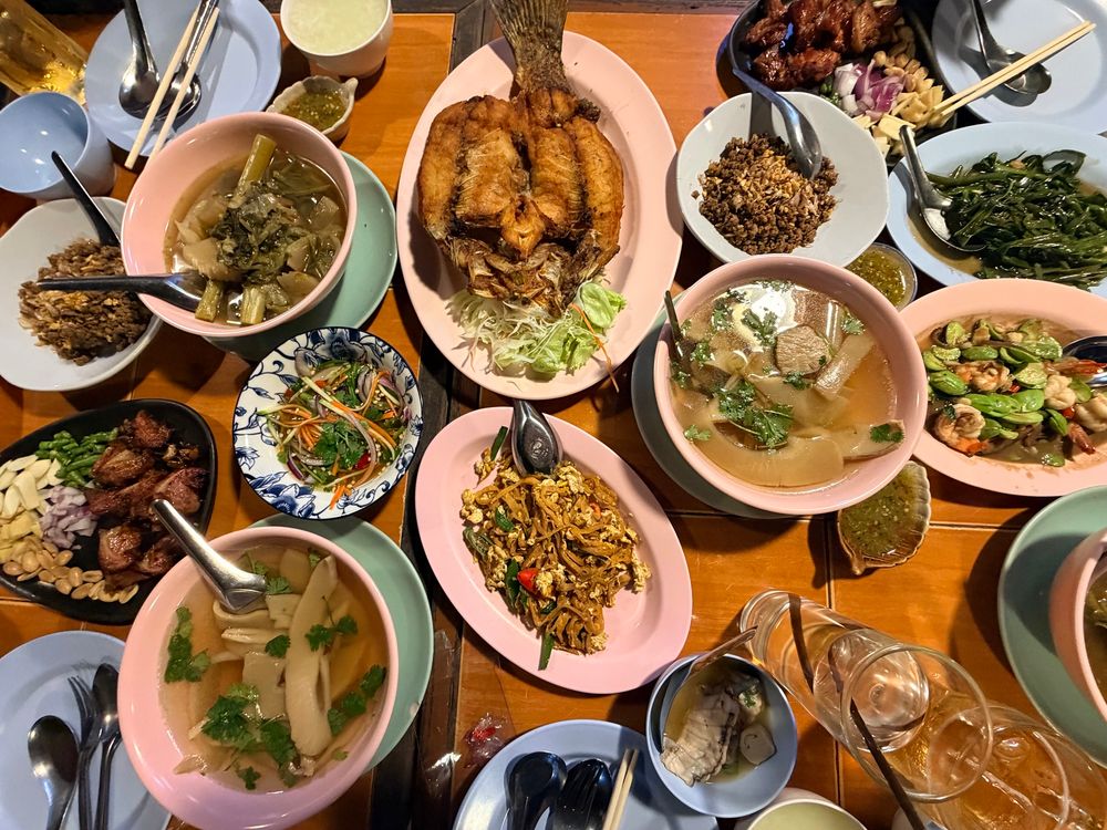 A table filled with a variety of Thai dishes, including fried fish, clear soups with vegetables, spicy stir-fries, a colorful papaya-style salad, sautéed greens, grilled meats served with fresh herbs and nuts, and assorted dipping sauces. The dishes are arranged in pink, blue, and patterned bowls and plates, creating a vibrant, abundant meal setting.