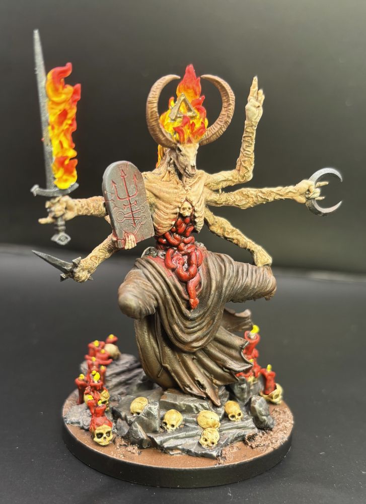 A side view of a painted model figure of a grotesque sorcerer with six arms, a goat skull face, and just all sorts of horror stuff going on.