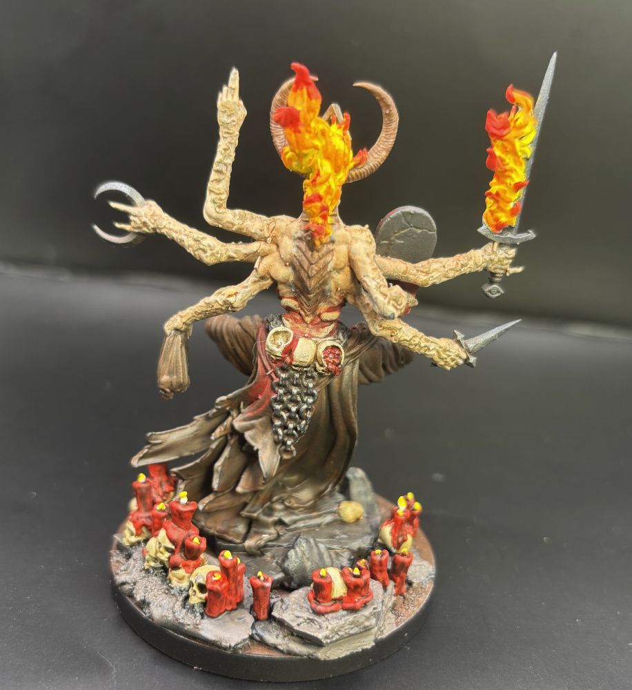 A rear view of a painted model figure of a grotesque sorcerer with six arms, a goat skull face, and just all sorts of horror stuff going on. Skulls and candles abound.