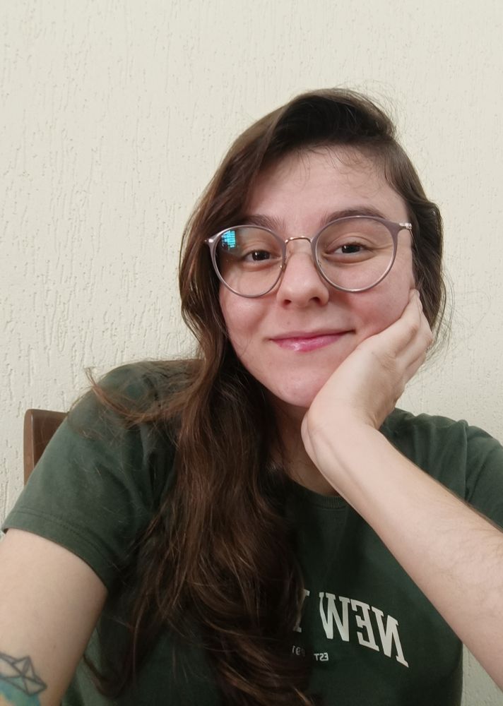 Selfie. Sou uma mulher branca, cabelos longos e ondulados de cor loiro escuro, uso óculos arredondados e estou com a mão direita apoiando o rosto, enquanto sorrio olhando a câmera. Uso uma camiseta verde escura e no meu braço esquerdo há uma tatuagem de barquinho de papel. Ao fundo, uma parede bege.