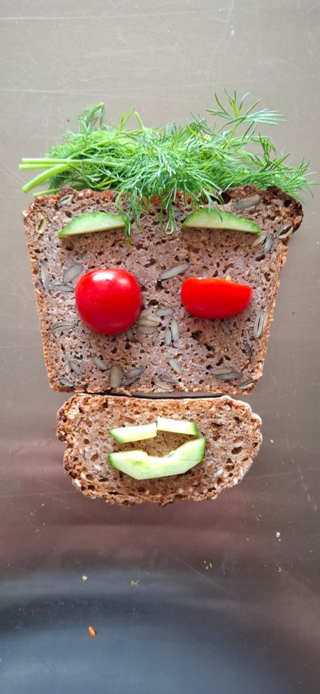 Ein Gesicht aus Brotscheiben. Kastenbrot oben, Baguette unten. Haare aus Dill. Augenbrauen aus Gurke, Tomaten als Augen, rechts halbiert wie zum Blinzeln.
Mund aus Gurke.