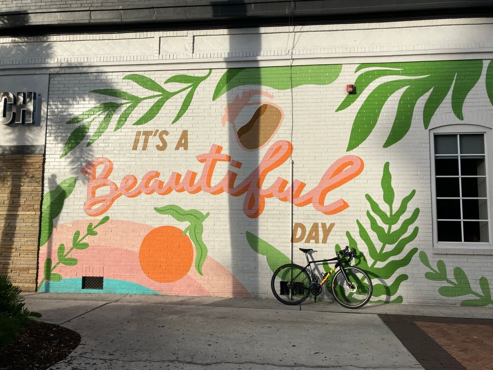 Black bike leaning against an orange citrus themed urban wall mural reading “it’s a beautiful day”