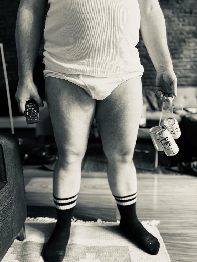 Black and white photo of a man from the waist down in white underwear and black socks, holding a TV remote and a six pack of beer