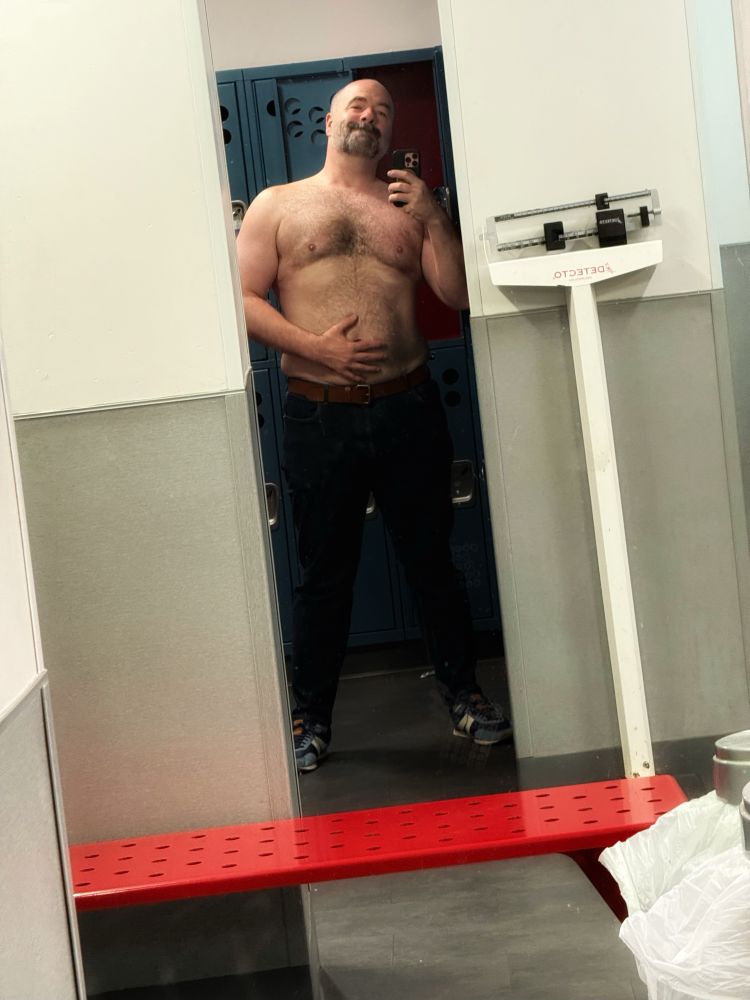 Distance selfie on the gym mirror. I’m shirtless with my hand on my belly, in dark jeans and a brown belt. Nodding to the camera.