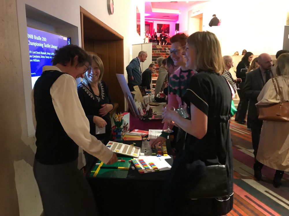Crowded room in the British Library Knowledge Centre. A row of tables along the left wall display braille Lego and historical braille artifices. People chat about the items in front of them. This was part of the drinks reception before a panel discussion. 