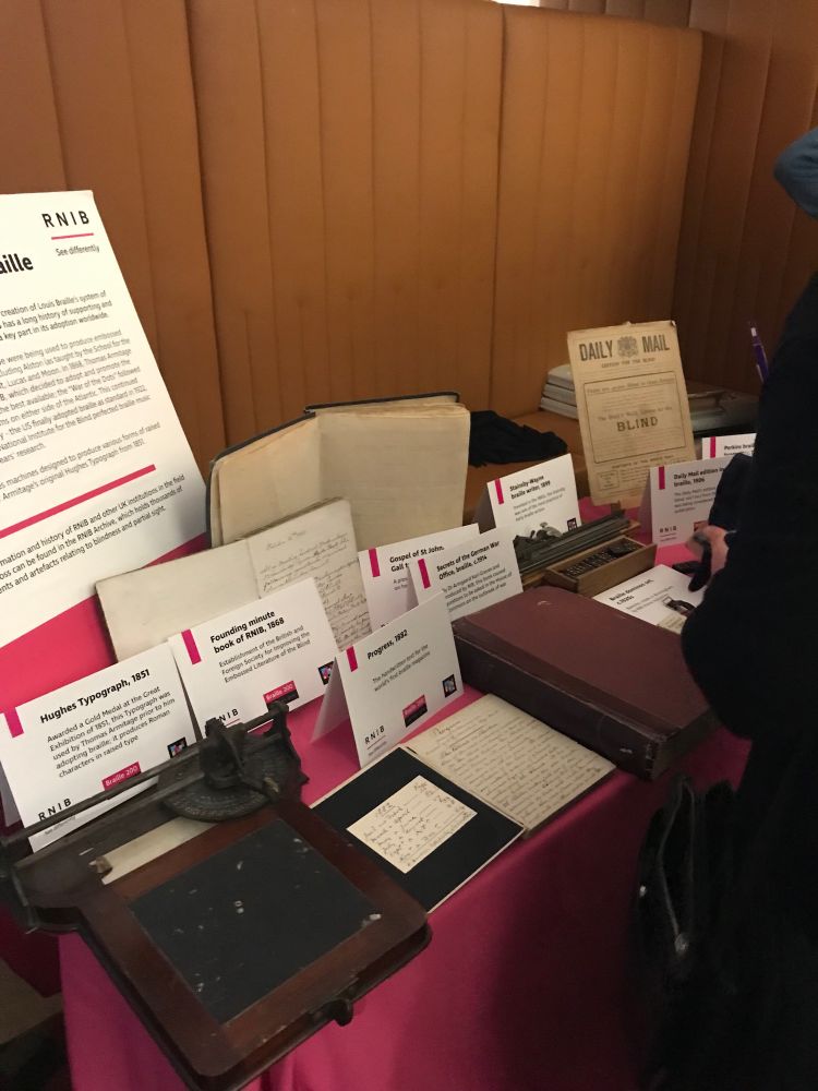 Table display of artefacts from the RNIB archive, as part of the drinks reception. Items include a Hughes Typograph, manuscript books of minutes and the text of Progress magazine, an embossed book on Gall type, a Braille copy of the Daily Mail, and a selection is writing tools and games. These sit on a pink table cloth, with captions printed in the RNIB font. 