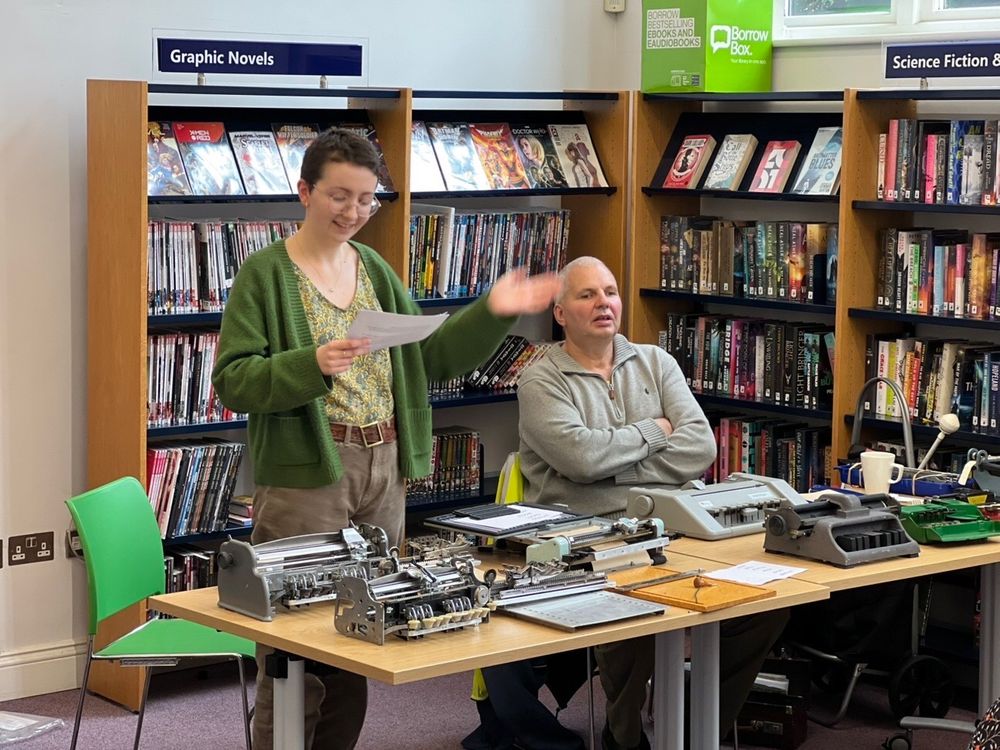 Tilly Guthrie (white woman, short dark hair, very cosy green cardigan) is gesticulating, mid talk, in front of an audience in Harrogate library. Co-speaker Alan is sitting next to her, behind a table covered in his collection of vintage braillers