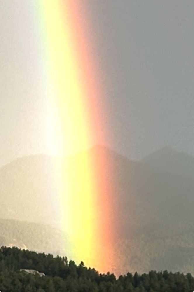 distinct vertical beams of a rainbow’s colored lights shoot upwards from tree covered hills dwarfing the outline of mountains behind them