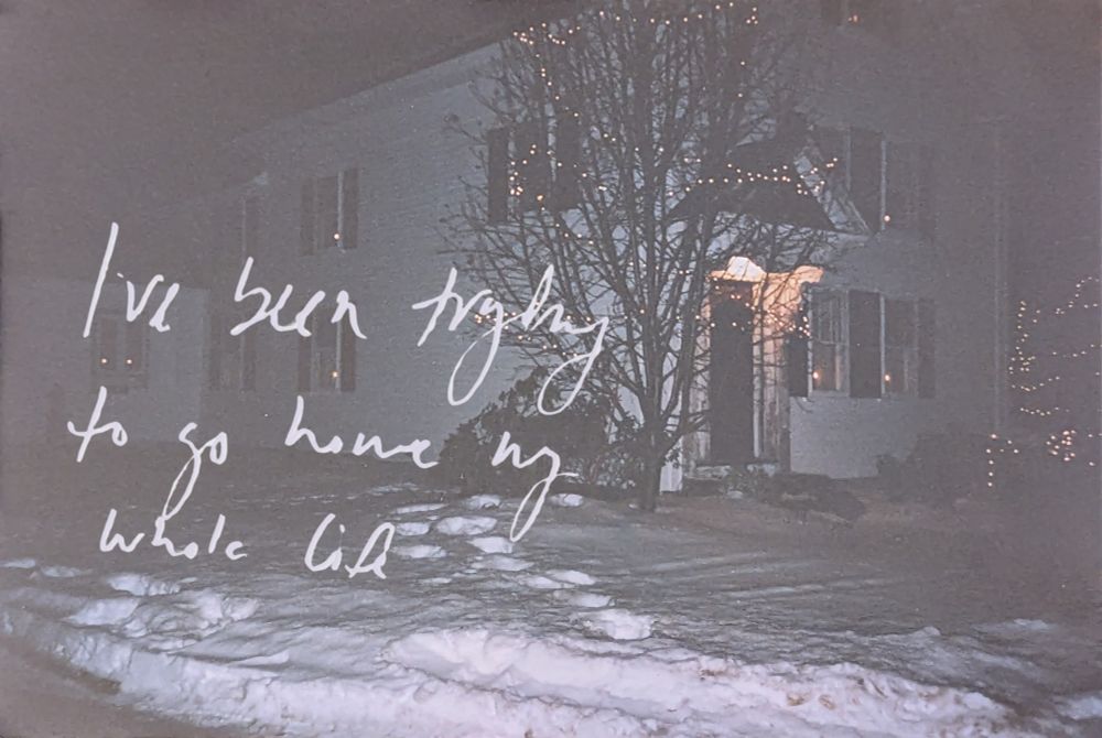 A grainy photo of a white house and a snowy yard, the photo is taken at night with flash. There are Christmas lights in the windows, as well as strung up on the trees and bushes out front. There are footprints in the snow leading towards the house. In white handwritten text reads "I've been trying to go home my whole life"