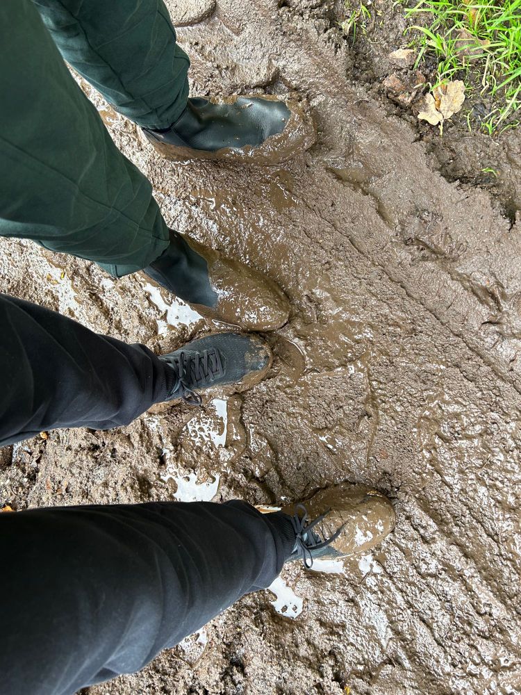 Looking down 2 sets of legs below thighs, one in black and the other in dark green joggers towards 2 pairs of very damp and muddy shoes, standing on a wet, muddy footpath.