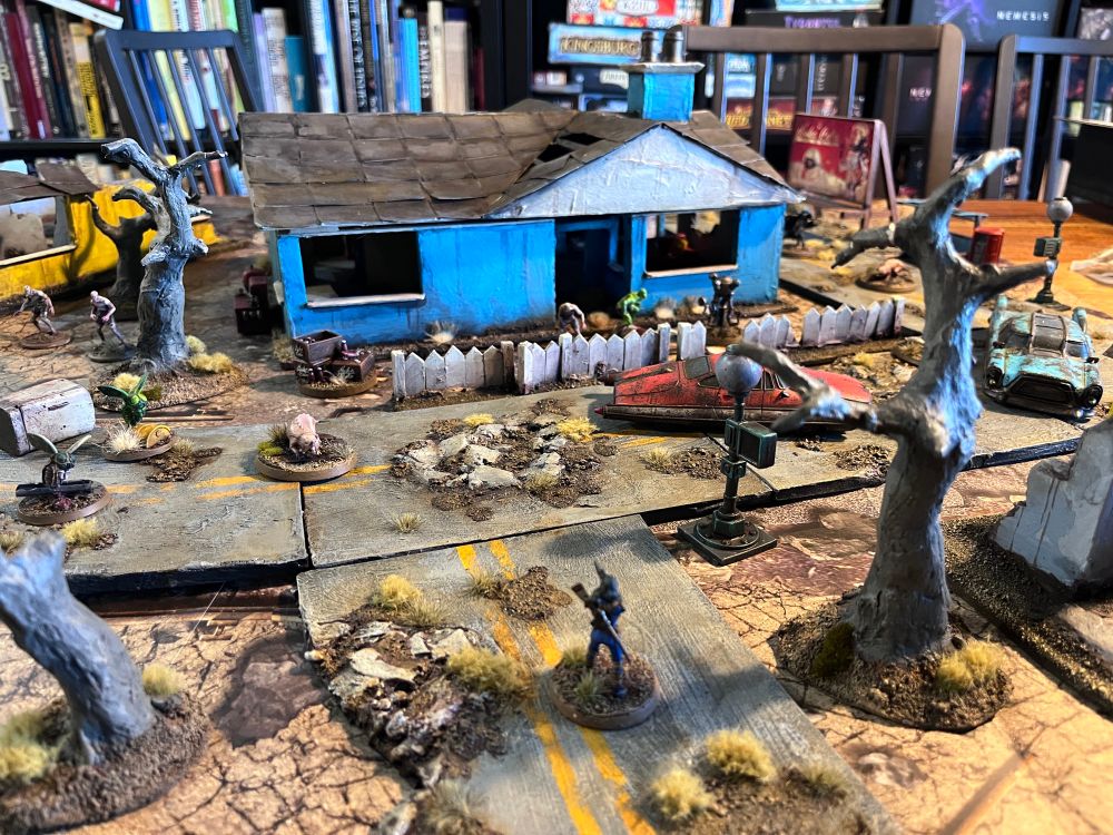 Fallout Wasteland Warfare miniatures game setup. Hand made terrain of Sanctuary Hills suburbia, with the Sole Survivor returning home after 220 years. Her new neighbours are bloat flies, rad roaches, ghouls and mole rats 