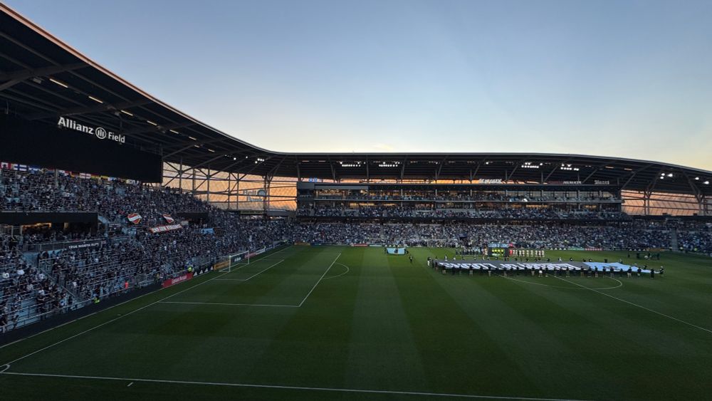 Wide-angle shot of the moment of silence with the “End Gun Violence” banner in the Wonderwall on the left and the traditional Loons banners and flags at midfield. The sun is setting orange behind Allianz.