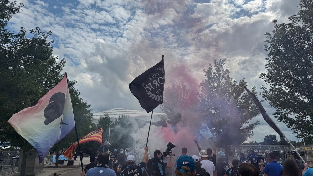 Marchers carry flags as smoke is deployed. Down a tree-lined street, Allianz Field stands in the distance. 