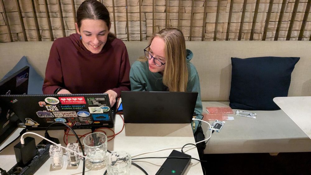 two people sitting at a table with two laptops, cables, and phones hacking together.