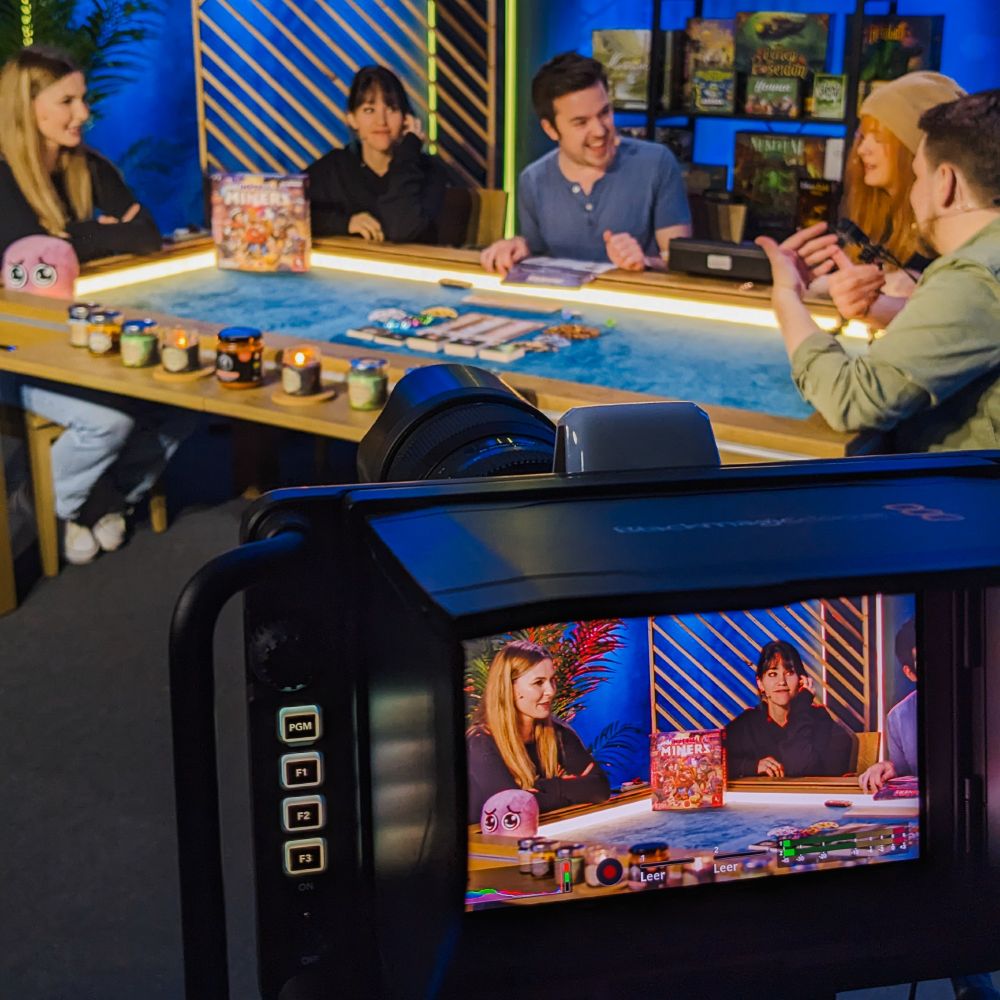 An Kameraausschnitt auf dem zu sehen ist: Fünf Personen spielen Brettspiele auf einem Tisch