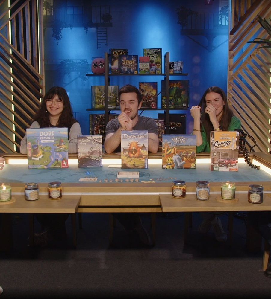 Minakeks, Daniel Staubesandt und Meloonie lachend an einem Brettspieltisch. Vor ihnen aufgestellt sind 5 verschiedene Brettspiele und Duftkerzen.