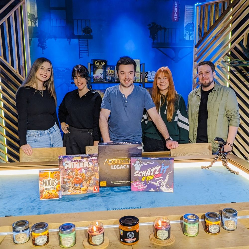 Fünf Personen vor einem großen Brettspieltisch. Auf dem Tisch befinden sich Spiele, Kerzen und ein Glas Löwenanteil. Vlnr Ninsi, Drachenkraut, Staubesandt, Froschmädchen und Loewepe