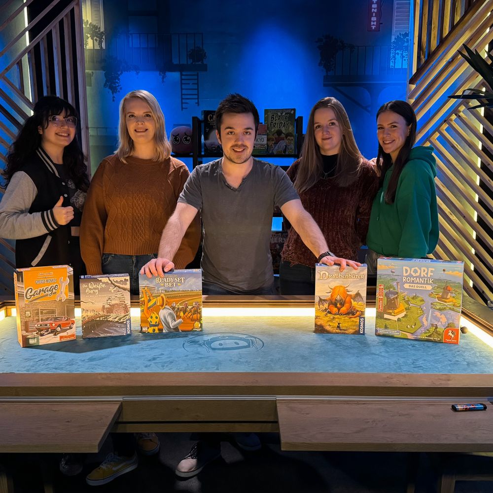 Fünf Personen vor einem großen Brettspieltisch. Auf dem Tisch befinden sich Spiele. Vlnr Minakeks, Lumina, Staubesandt, Raya und Meloonie