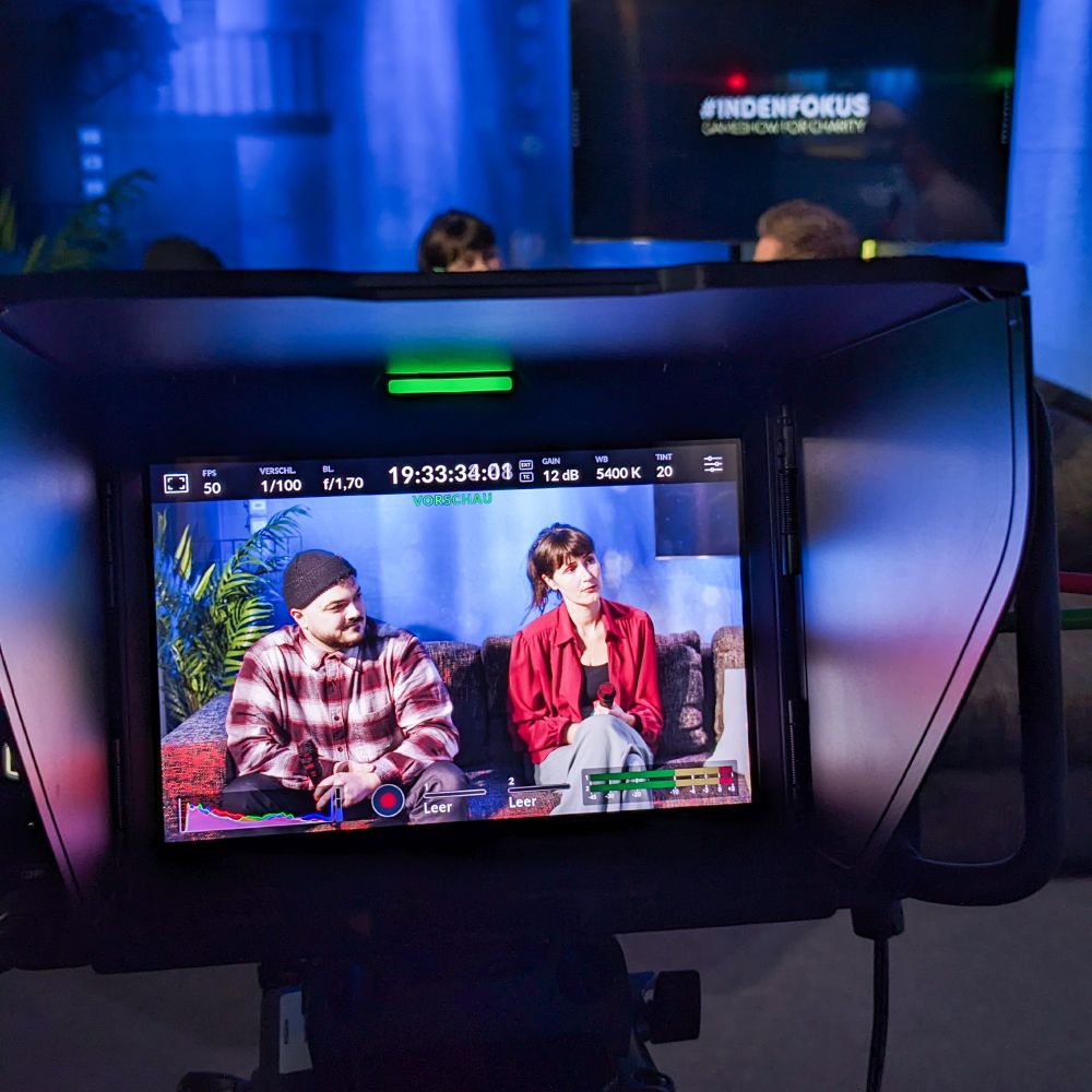 Ein abfotografierter Kamerabildschirm, auf dem zwei Menschen in einem Interview auf der Couch zu sehen sind.