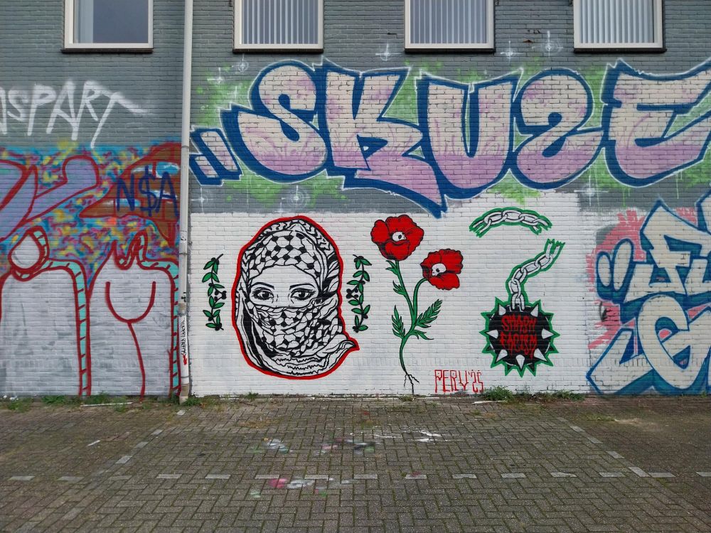 Een foto van een muur van een kantoorpand, op een verlaten parkeerplaats. Op de muur is graffiti te zien. Veel persoonlijke graffiti en kunst, maar voornamelijk, een afbeelding van een vrouw met een Kufiyeh over haar hoofd. Het hoofd is rood omlijnd. Ernaast twee olijftakken, en een klaproos met twee bloemen, en daarnaast een strijdvlegel met een gebroken tekst. Een strijdvlegel is een bal met punten aan een ketting. Op de bal staat de tekst 'Smash Fascism'. Eronder een persoonlijke tag, Pery '25. 