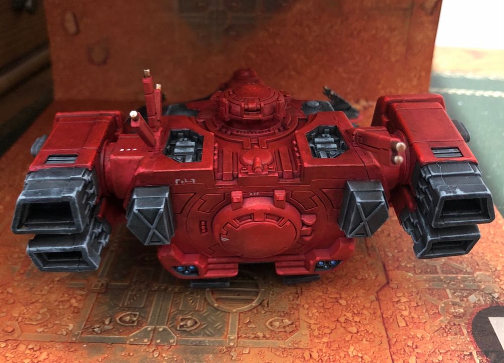 Back side of a T’au Devilfish transport miniature, painted in reds and blacks with gold, white, and blue details, over a sand and rust background