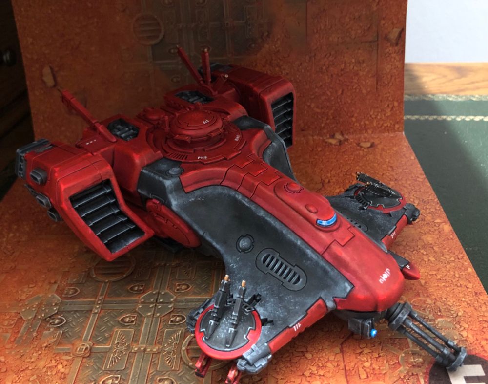 Right side of a T’au Devilfish transport miniature, painted in reds and blacks with gold, white, and blue details, over a sand and rust background