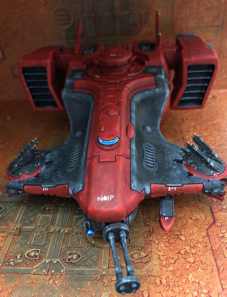 Front side of a T’au Devilfish transport miniature, painted in reds and blacks with gold, white, and blue details, over a sand and rust background