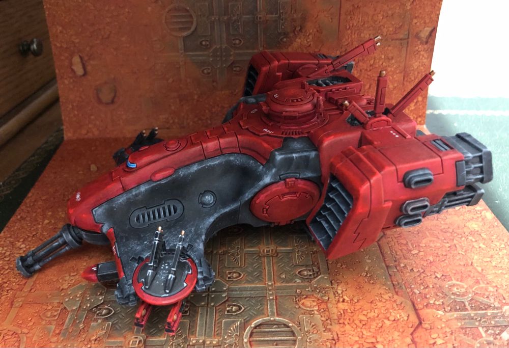 Left side of a T’au Devilfish transport miniature, painted in reds and blacks with gold, white, and blue details, over a sand and rust background